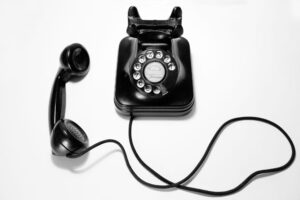 Black vintage rotary dial telephone with the handset off the hook, lying to the left of the base, connected by a coiled cord on a white background.