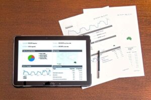 A tablet displaying graphs and analytics is placed on a wooden desk, surrounded by papers with more charts and a pen.