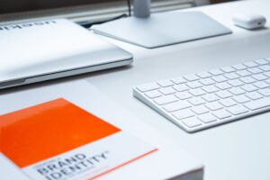 A modern desk setup featuring a closed laptop, a white keyboard, a monitor, and a book titled "brand identity" with an orange cover, all under soft lighting.