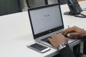 A person's hands typing on a laptop with the google homepage displayed on the screen, set on a desk next to a smartphone and a desk phone.