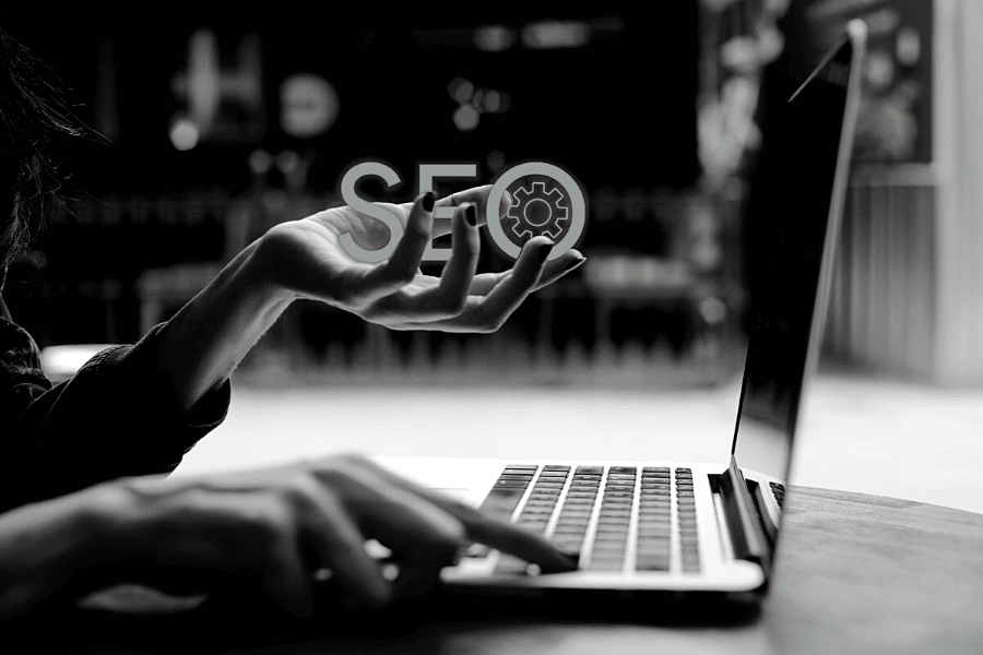 A person sits at a laptop in a dimly lit room, holding a translucent, floating "seo" and gear icon in one hand, symbolizing search engine optimization work.