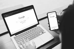 A grayscale image depicting a person using google on both a laptop and a smartphone, with the screens visible and aligned centrally on the devices.