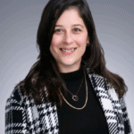 Woman with long dark hair in a black turtleneck and plaid coat, smiling at the camera.