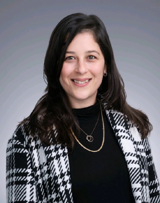 Woman with long dark hair in a black turtleneck and plaid coat, smiling at the camera.