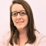 Woman with long brown hair, glasses, and a light shirt smiling against a plain background.
