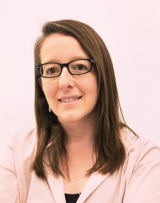 Woman with long brown hair, glasses, and a light shirt smiling against a plain background.