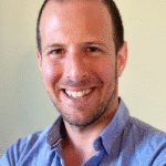 Smiling man in a blue shirt, facing the camera with a light-colored background.