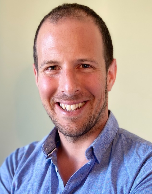 Smiling man in a blue shirt, facing the camera with a light-colored background.