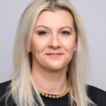 Woman with straight blonde hair wearing a black blazer and a gold beaded necklace, smiling.