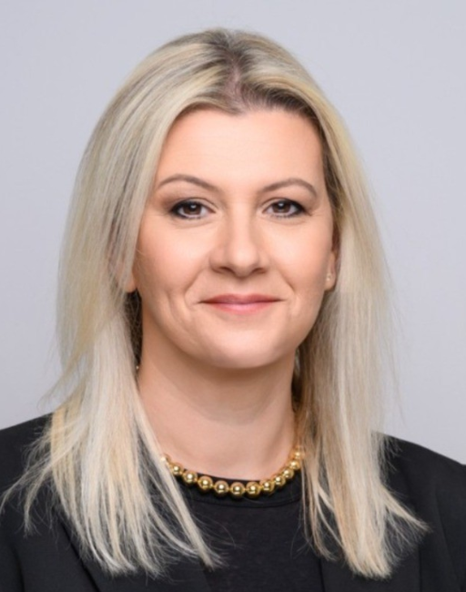 Woman with straight blonde hair wearing a black blazer and a gold beaded necklace, smiling.