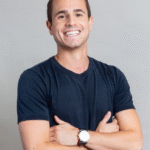 Smiling man in a navy t-shirt with arms crossed, standing against a light gray background.