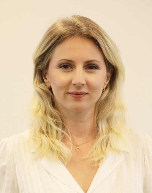 Woman with blonde hair wearing a white blouse, smiling softly against a plain background.