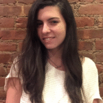 Woman with long dark hair smiling in front of a brick wall, wearing a white top.