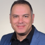 Smiling man in a blue blazer and black shirt against a plain light background.