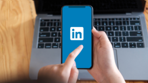 Person holding a phone with LinkedIn Posting open, in front of a laptop on a desk.