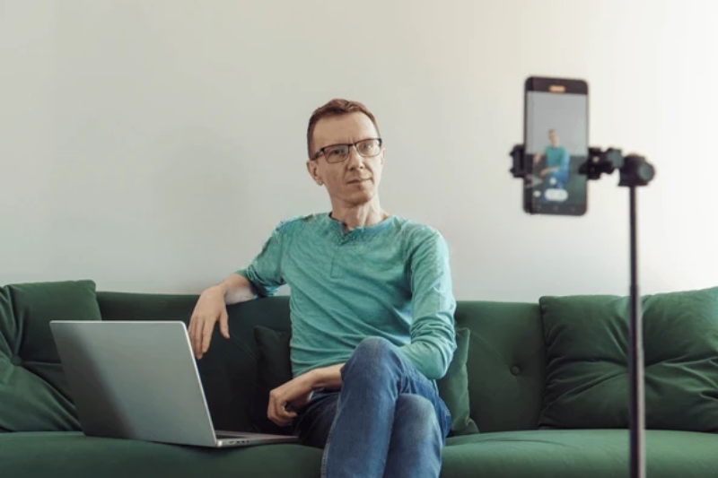Man with glasses on green sofa, laptop on lap, recording video marketing content on tripod.